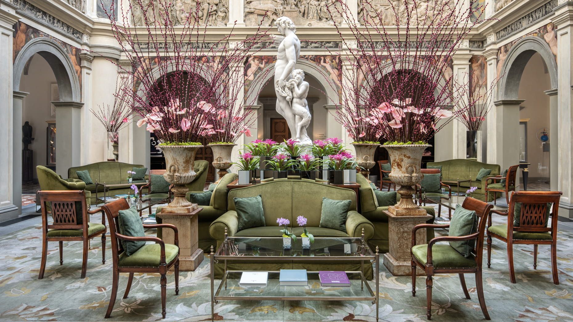 Elegant seating area in a courtyard. Green sofas and chairs, flowers, and a statue, surrounded by archways. Hotel Portraits –Four Seasons Hotel Firenze