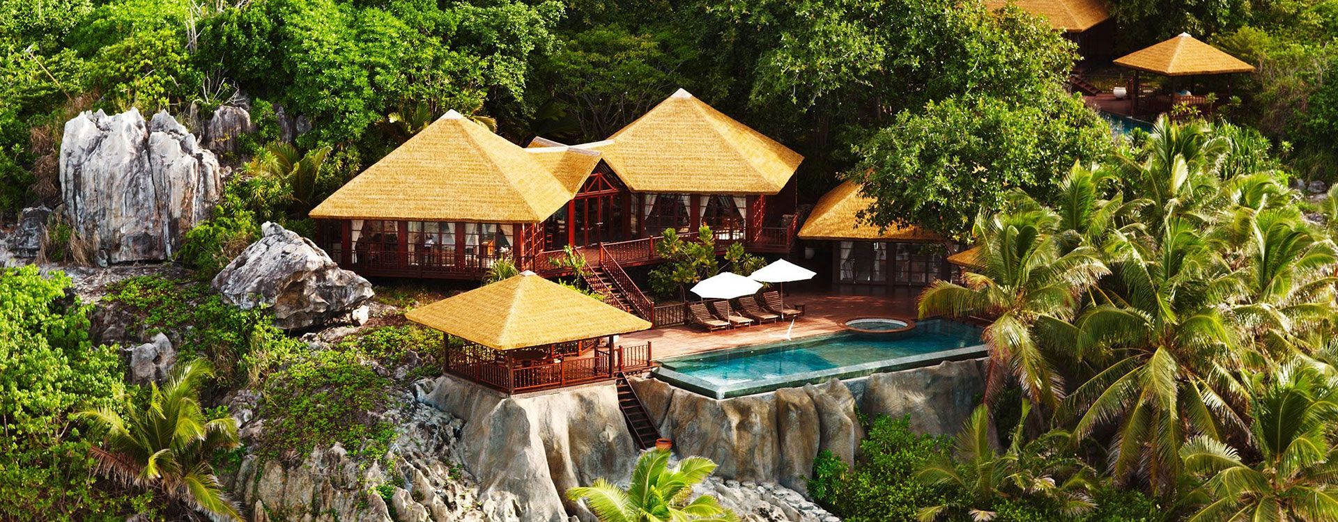 Tropical resort with thatched-roof structures, a pool, and lush greenery on a cliffside. Fregate Island Private, Seychelles
