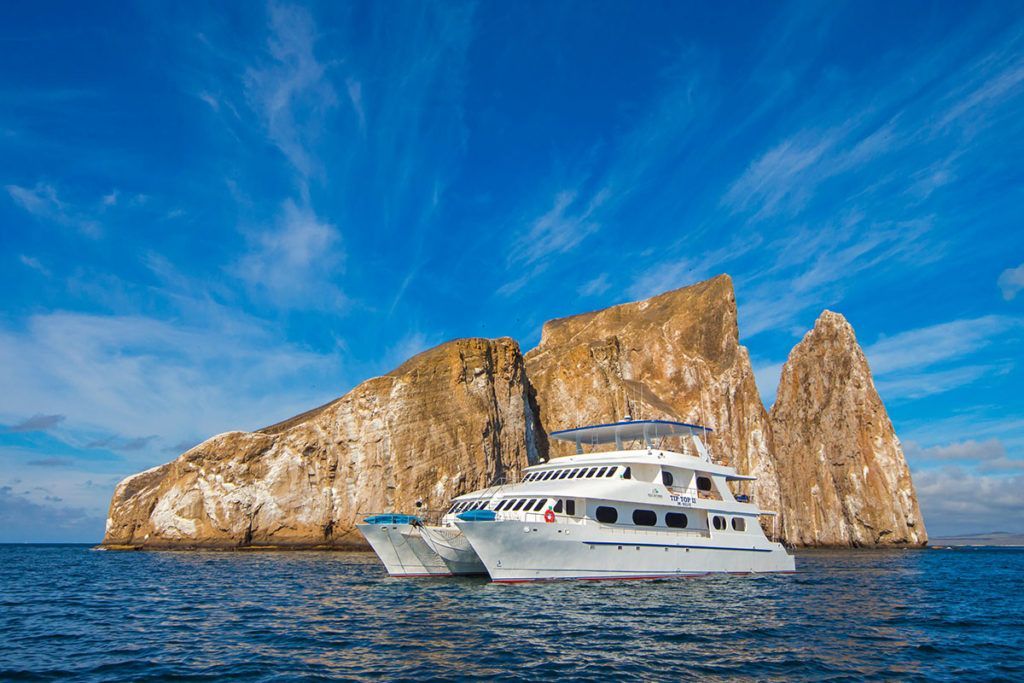 Catamaran boat in front of rugged, rocky island in blue ocean, under a bright blue sky.