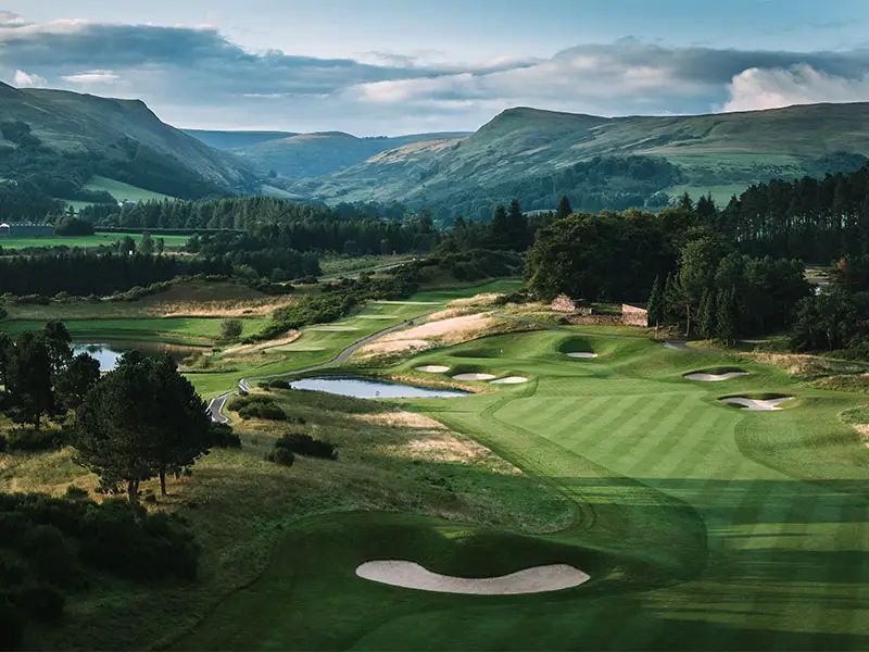 Golf course with green fairways, bunkers, and pond, nestled in a valley with mountains in the background. Gleneagles