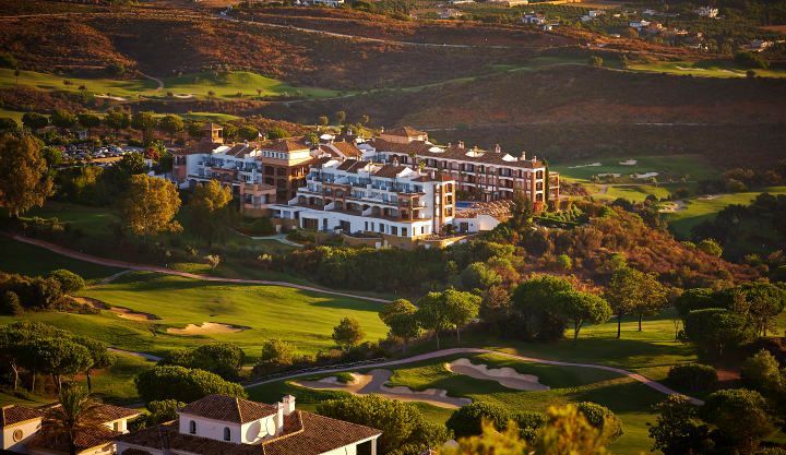 Luxury resort nestled in a green valley, with golf course and buildings with white walls and brown roofs.