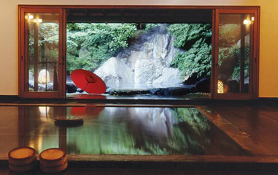 A Japanese-style bathhouse with a sliding glass door open to a natural waterfall. A red umbrella sits by the water. Gora Kadan