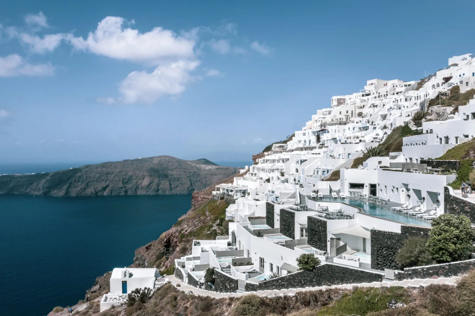 White buildings cascading down a cliffside in Santorini, Greece, overlooking the blue Aegean Sea. Grace Hotel Santorini, Auberge Resorts Collection