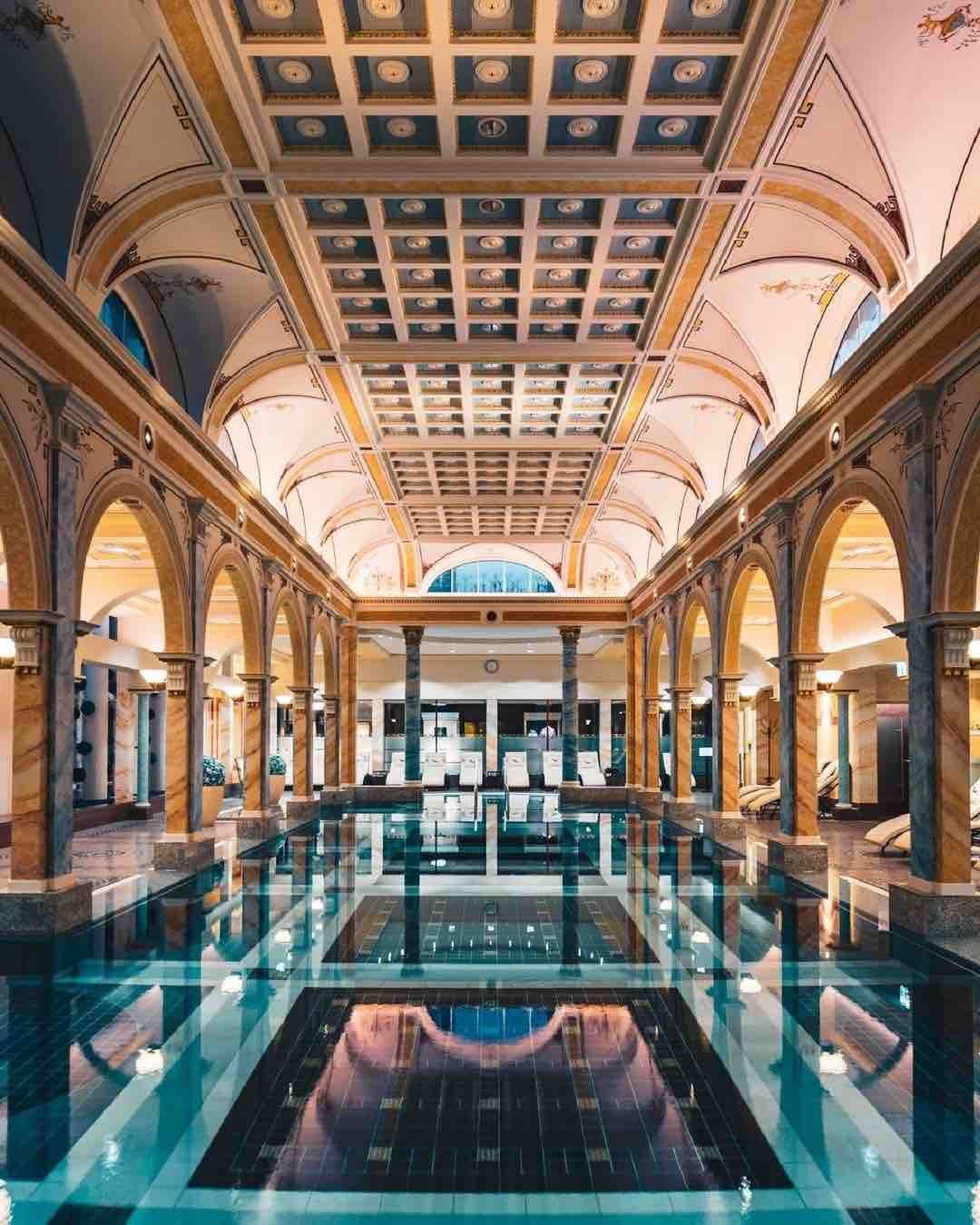 Indoor swimming pool with ornate arched columns and a patterned ceiling, reflections in water.