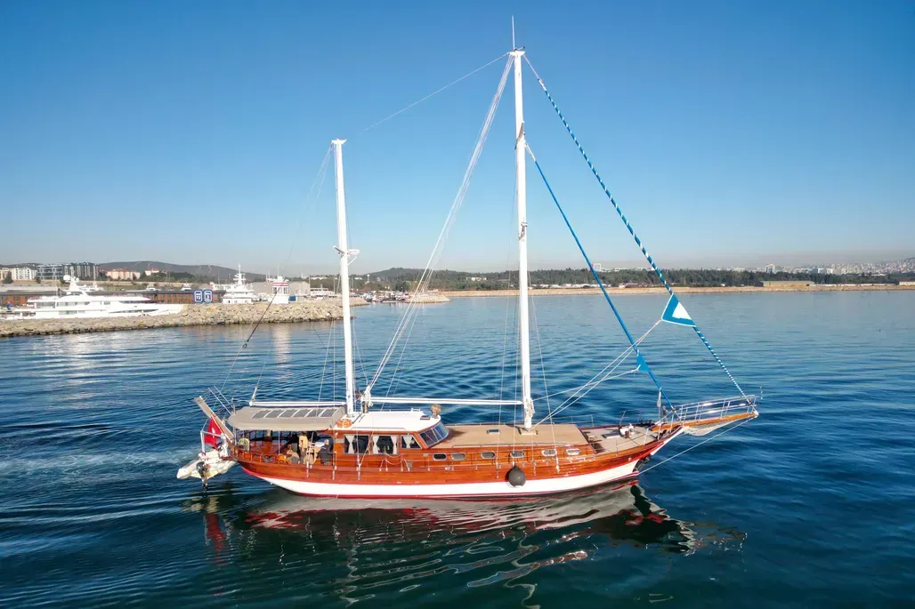 Wooden sailboat on blue water with a harbor in the background under a clear sky. Gulets & Traditional Wooden Vessels