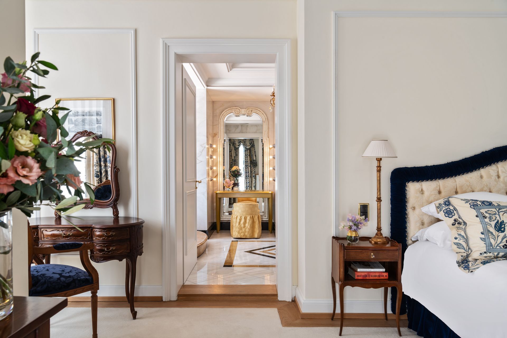 Bedroom with a vanity and a glimpse into a bathroom; neutral walls, wooden furniture, and a blue upholstered headboard.