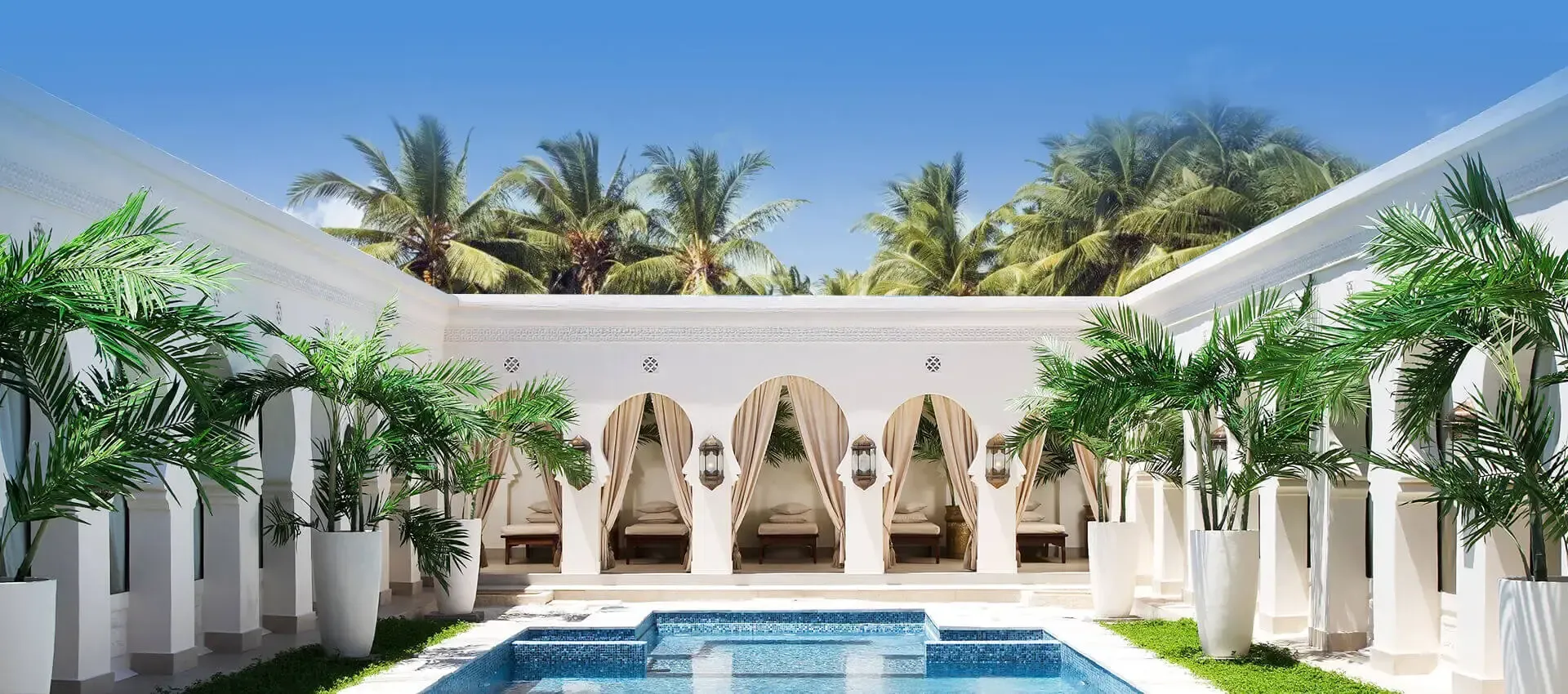 A courtyard with a swimming pool, flanked by white colonnades and palm trees, leading to curtained cabanas.