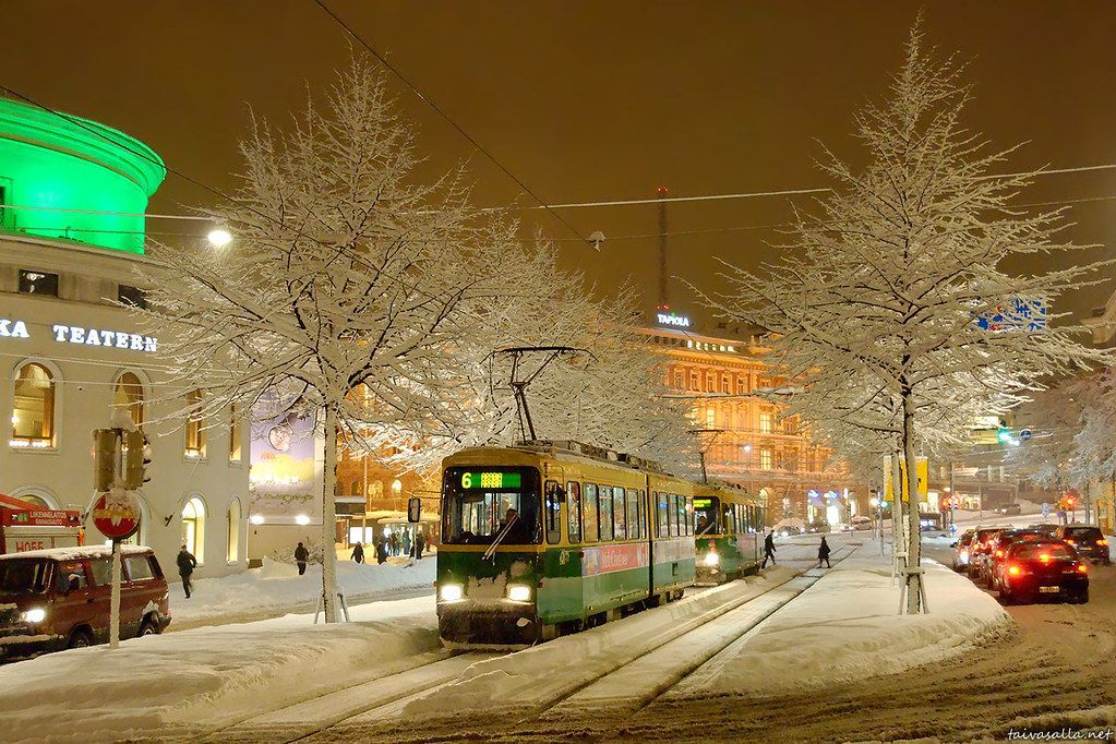 A green tram travels down a snowy street lined with snow-covered trees and buildings at night. Helsinki