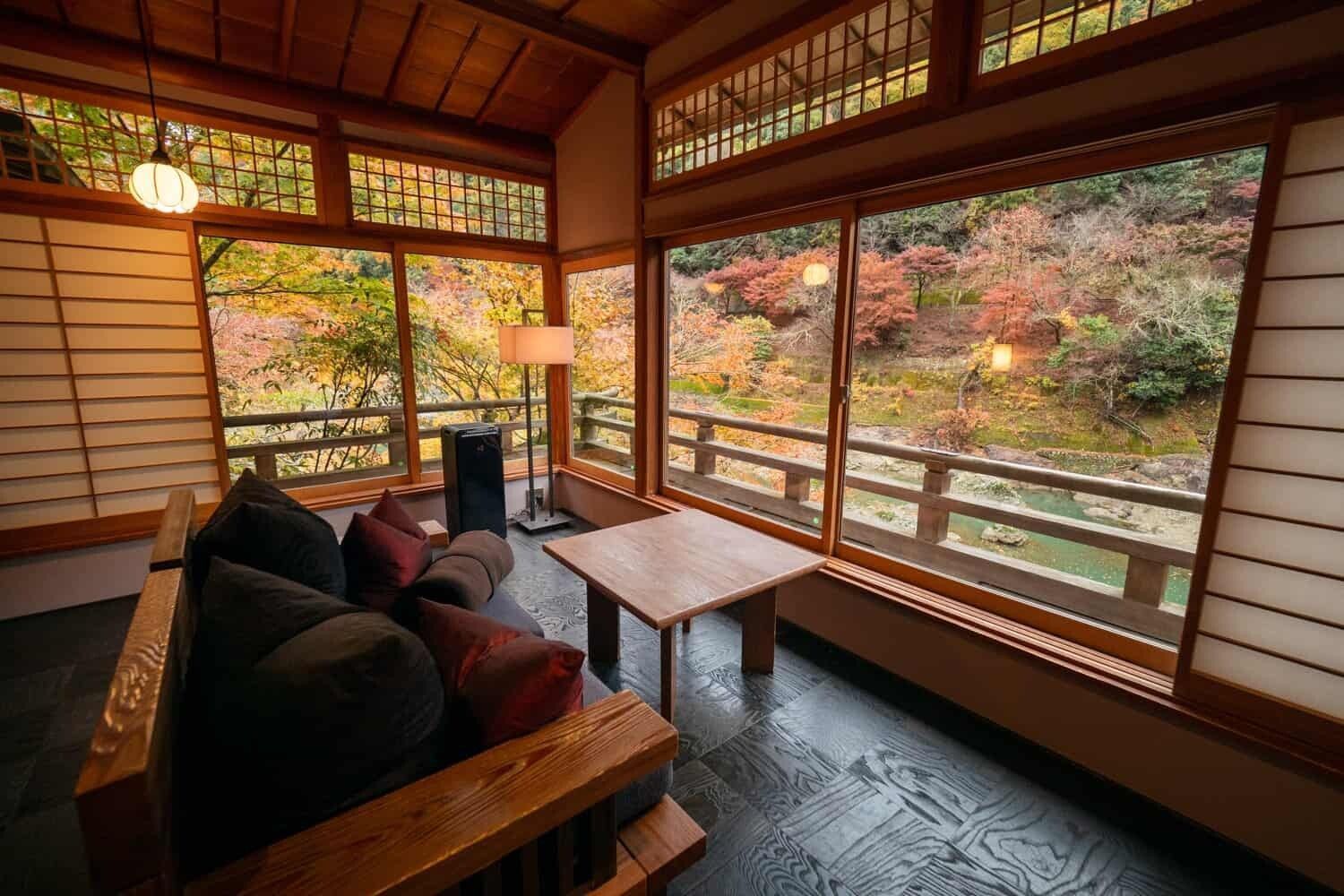 A cozy room with dark cushions, a wooden table, and large windows overlooking a colorful autumn forest. HOSHINOYA