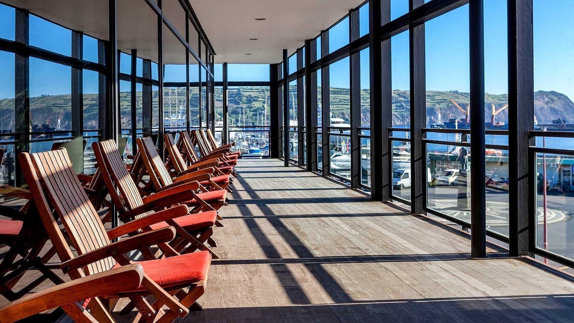 A row of red-cushioned wooden chairs on a sunlit indoor deck overlooking a harbor and rolling hills.