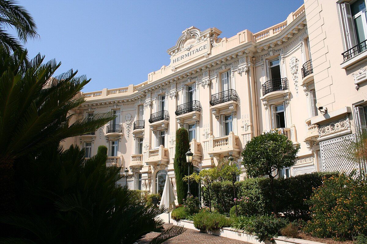 Hotel Hermitage Monte-Carlo Beige ornate building with balconies and a sign at the top; lush green landscaping in front of the building.