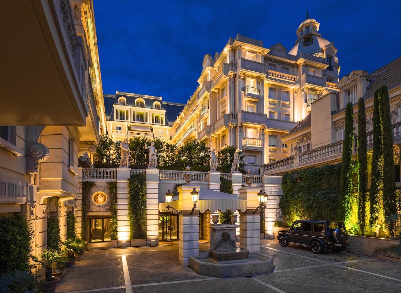 Hotel Metropole Monte-Carlo Exterior view of a grand, illuminated hotel at dusk. The building has a classical design with ornate details and a courtyard entrance.