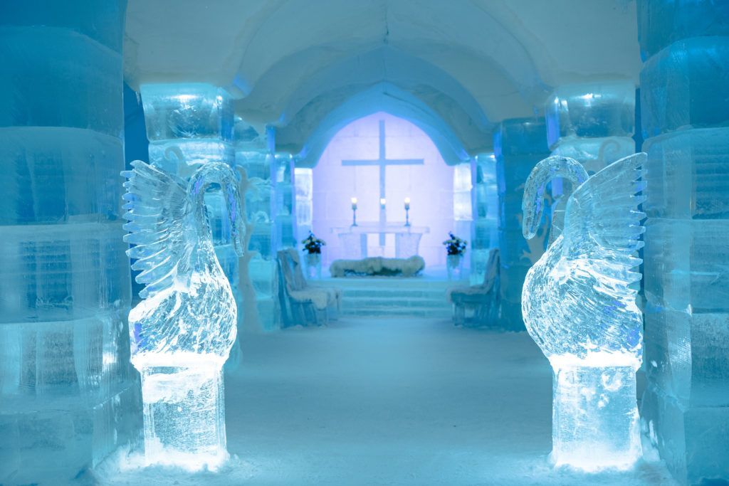 A chapel made entirely of ice, featuring translucent bird sculptures in the foreground and an illuminated cross altar. Sorrisniva, Alta
