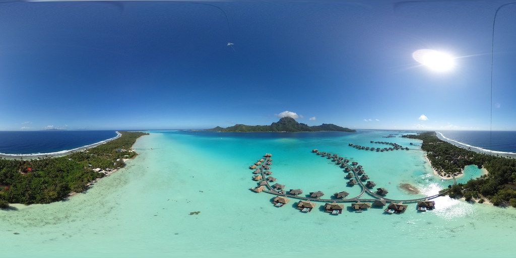 InterContinental Bora Bora Resort & Thalasso Spa Overwater bungalows on a turquoise lagoon with a mountainous island in the background, under a bright blue sky.
