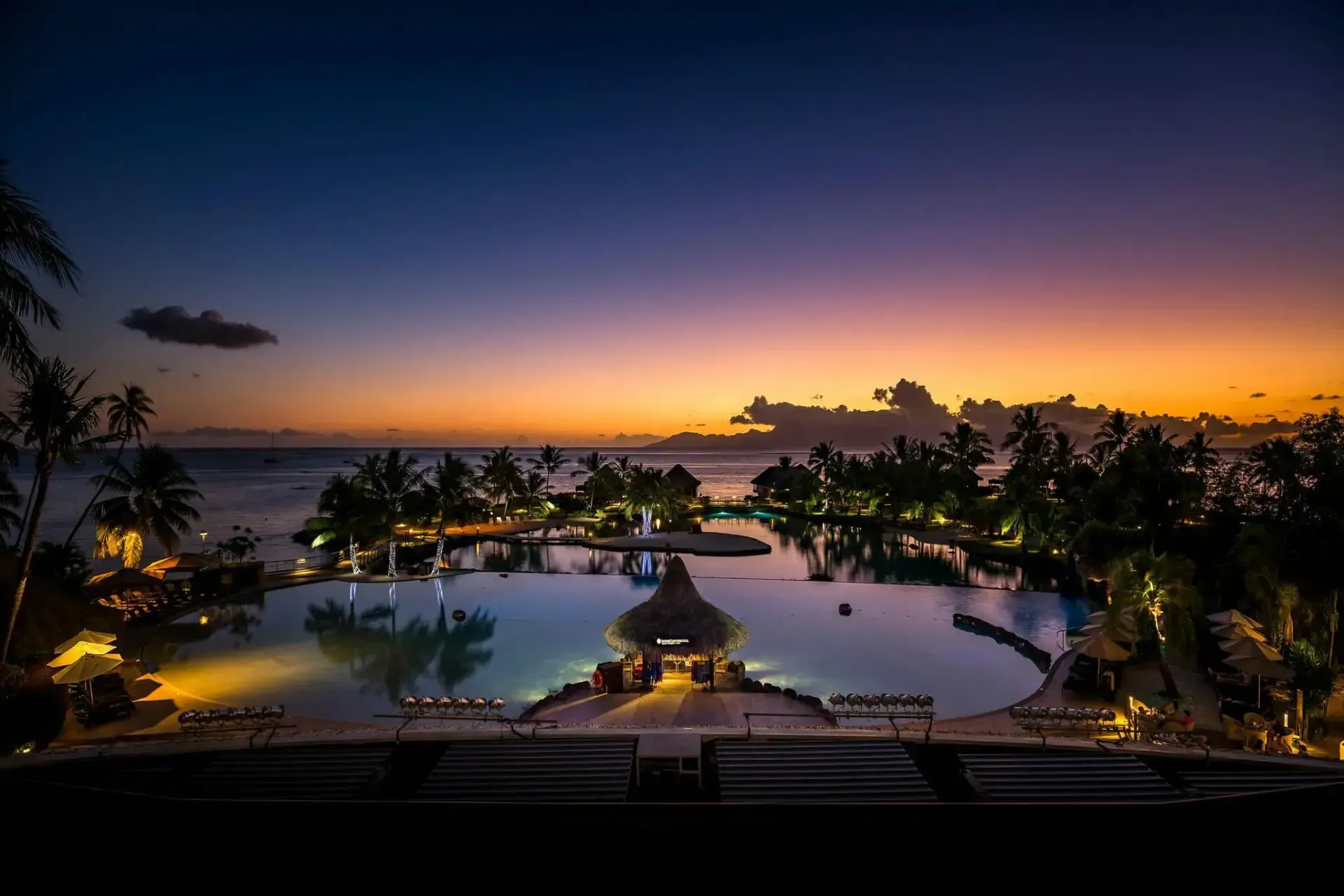 A tropical resort swimming pool glows with warm lights at sunset, framed by palm trees against a purple and orange sky. InterContinental, Tahiti
