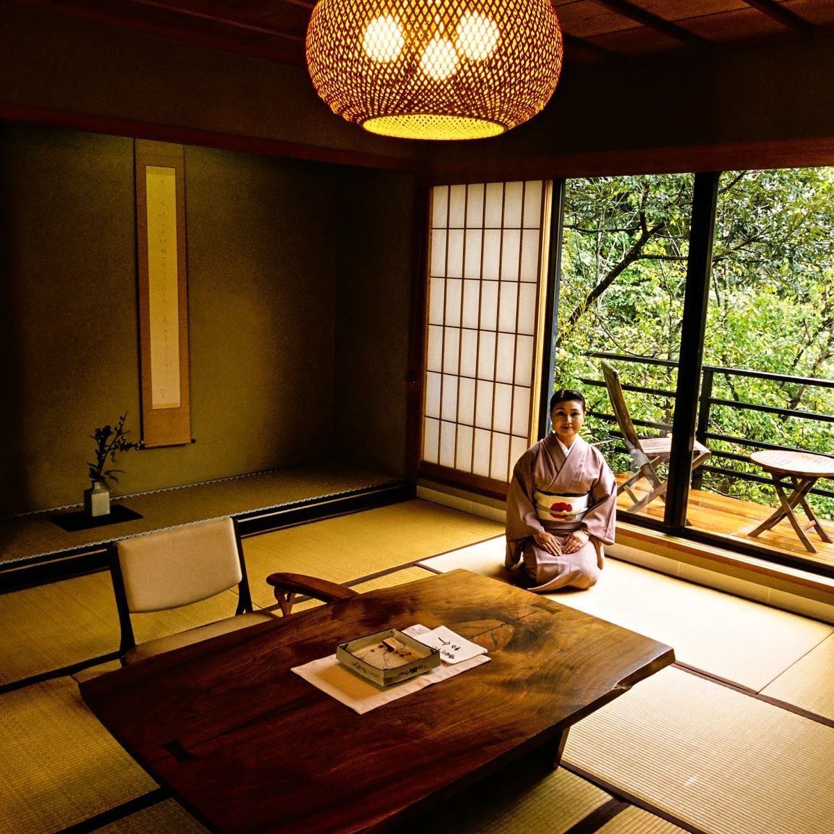 Woman in kimono kneels in Japanese-style room with forest view. Dark wood table, woven light fixture, tatami mats.