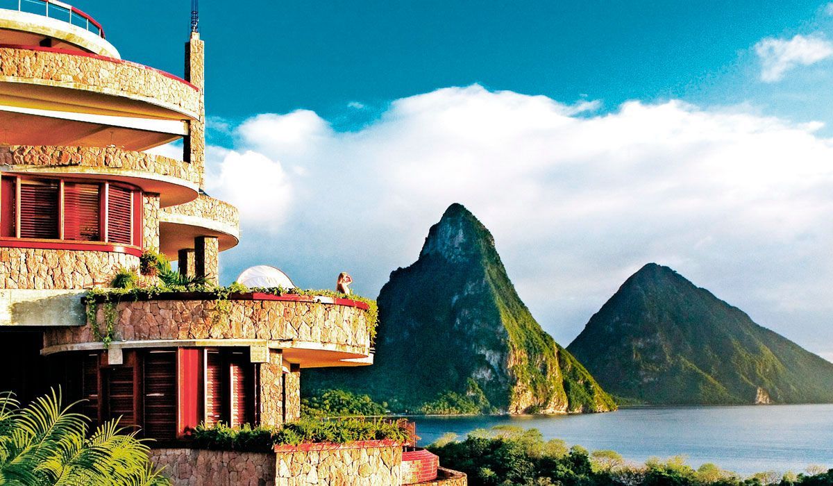 A tiered stone building overlooks the Caribbean Sea with the iconic Piton mountains rising in the distance under a blue sky. Jade Mountain, St Lucia