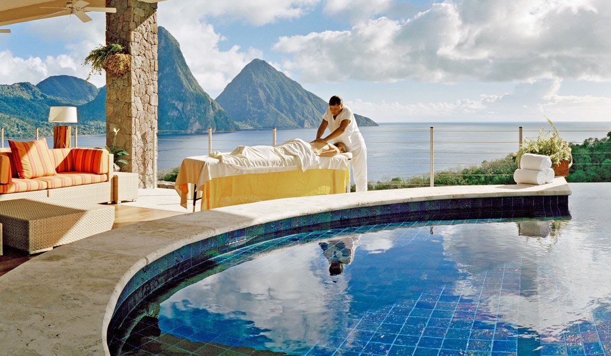 A massage therapist performs a treatment on a client at an outdoor spa overlooking a tropical bay and two mountain peaks. Jade Mountain, St Lucia