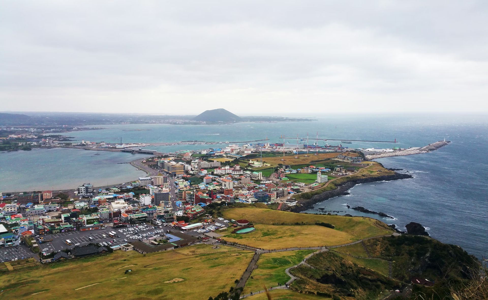 Coastal town with colorful buildings, a harbor, and grassy hills under a cloudy sky Jeju