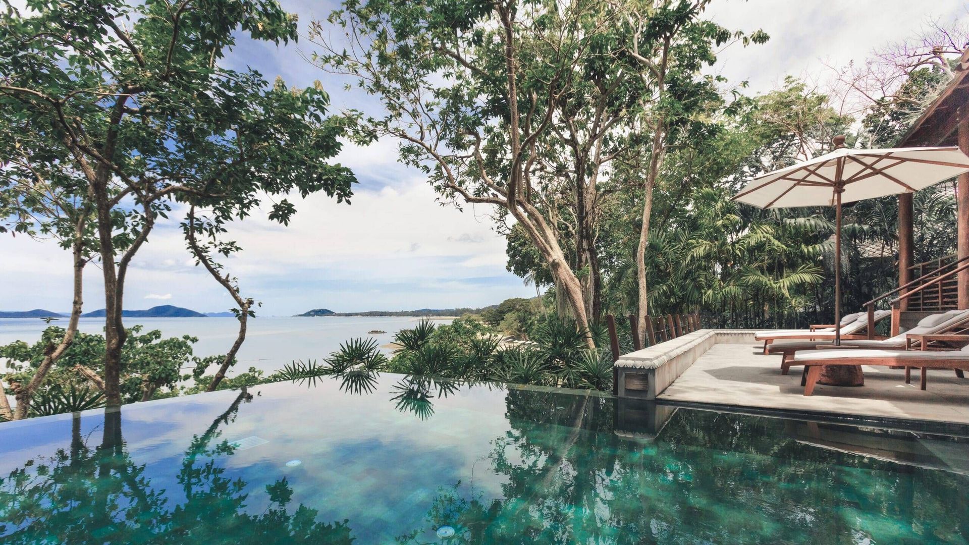 An infinity pool overlooking a tropical bay with lounge chairs and an umbrella on a wooden deck surrounded by trees. Kamalaya, Koh Samui
