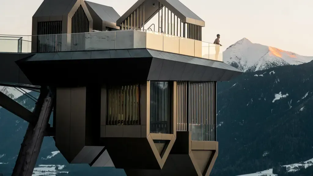 Modern geometric building perched on a mountainside, with person on balcony, snowy peaks in the background. Kronoplatz