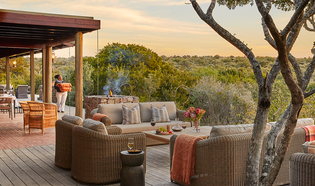 Outdoor lounge with seating, a fire pit, and a person carrying a basket, overlooking a savanna at sunset. Kwandwe Ecca Lodge (Eastern Cape)