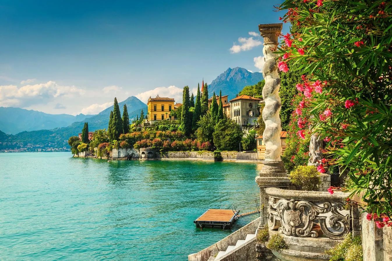 Villas along the tree-lined shore of Lake Como, Italy, viewed from a stone terrace with a decorative pillar and flowers.