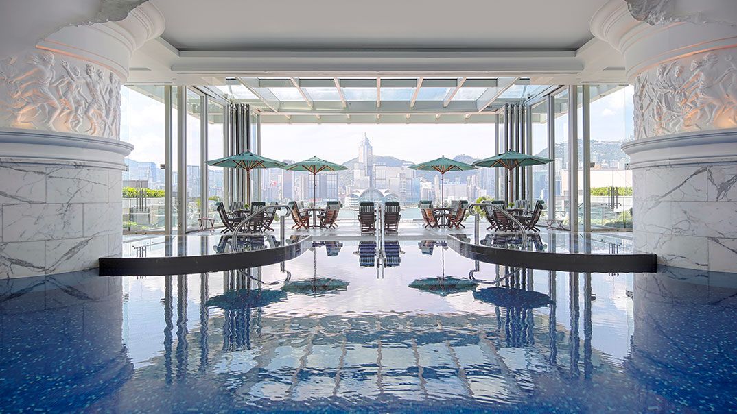 A luxurious pool reflects an open-air dining area with city views. Blue water, marble columns, tables, and umbrellas.  The Langham, Hong Kong
