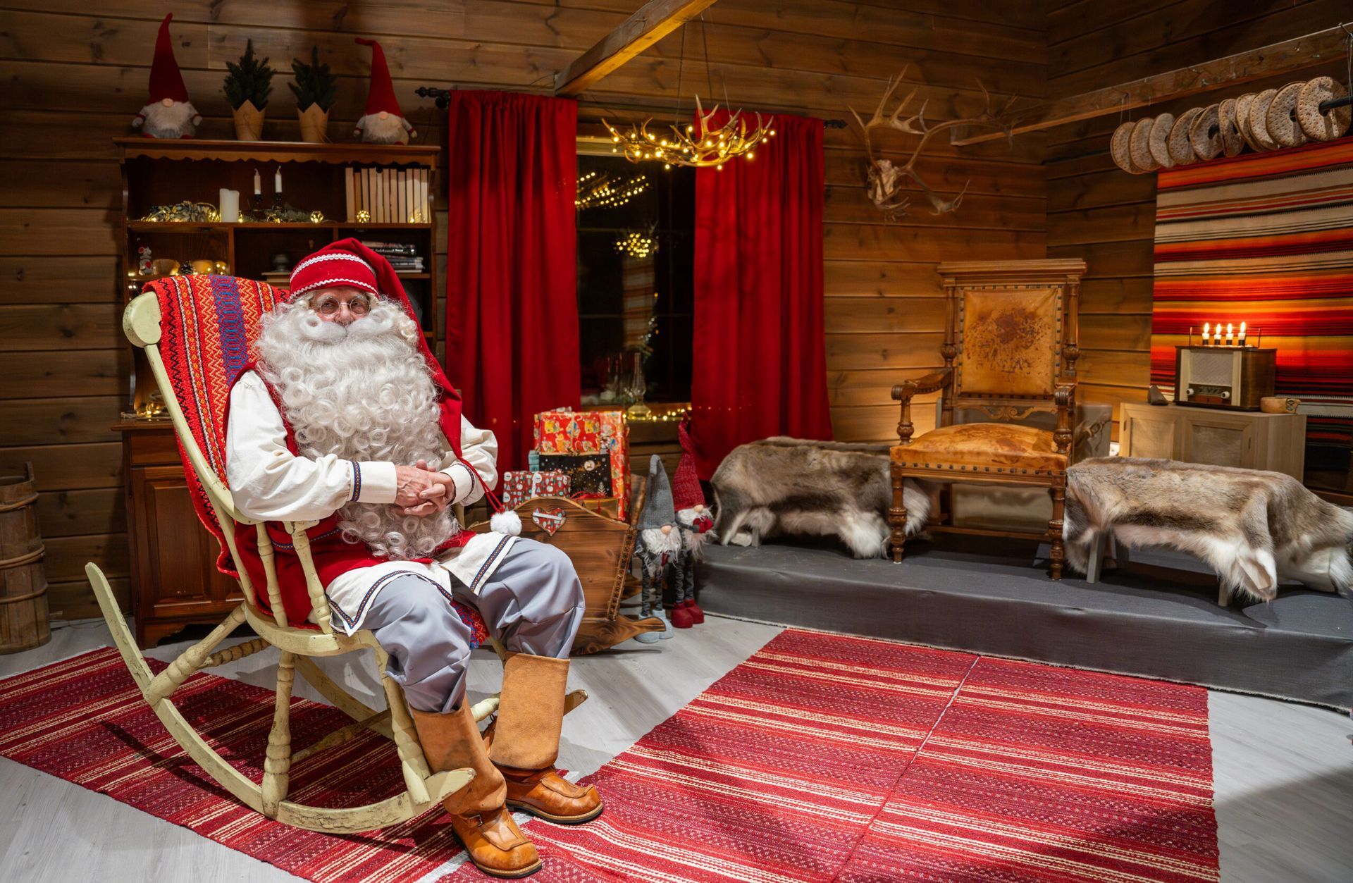 Santa Claus in rocking chair, inside cozy cabin with gifts and reindeer hides.