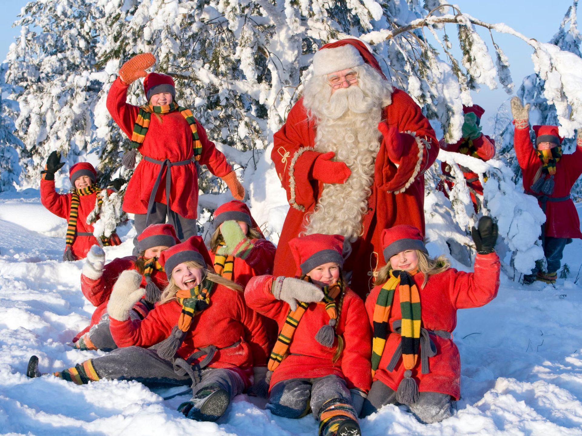 Santa Claus surrounded by elves in a snowy forest. They wear red outfits and scarves, waving and smiling.