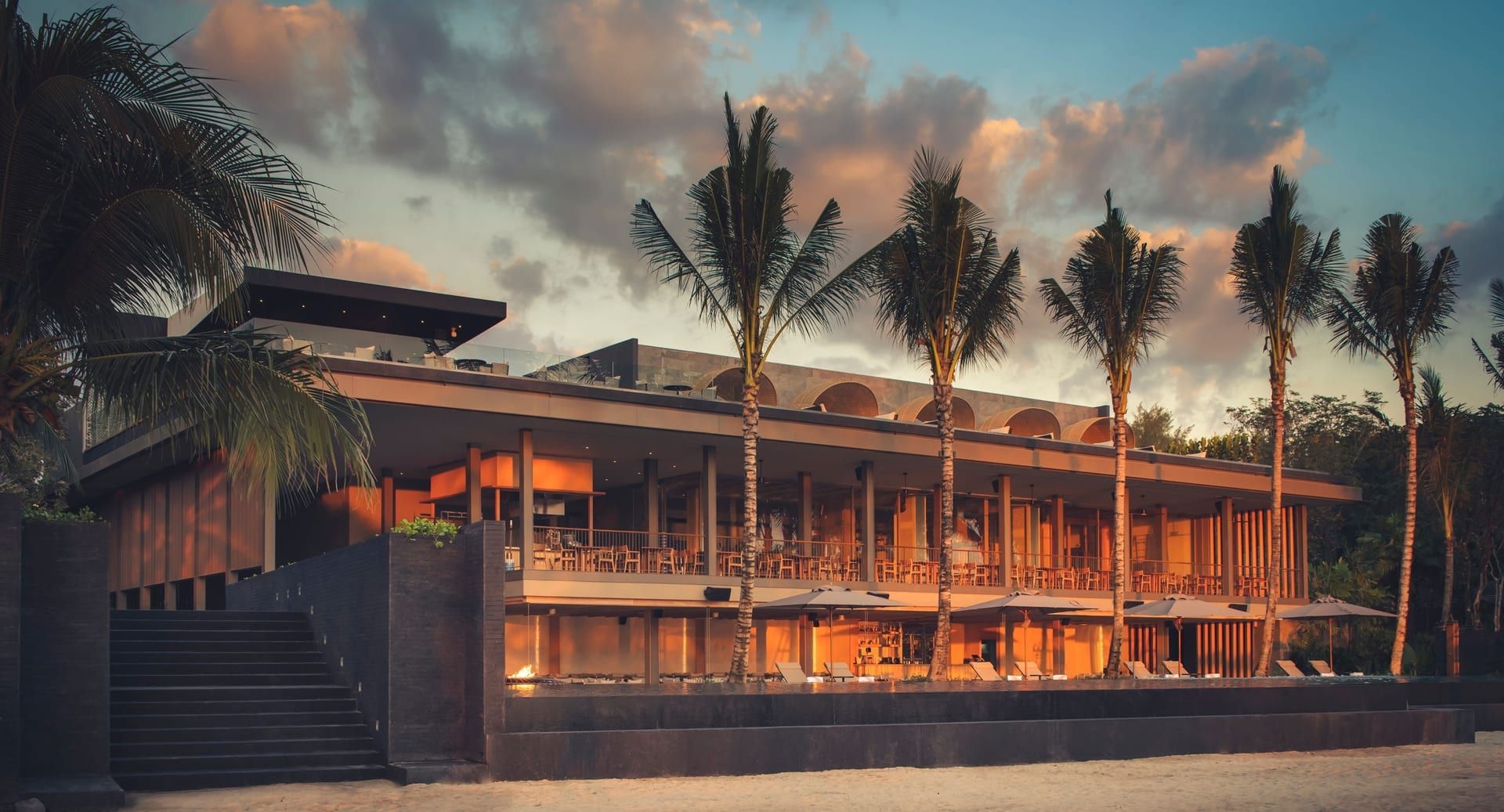 A modern beachside building with a long, open veranda lit by warm light at dusk, with palm trees. Layan