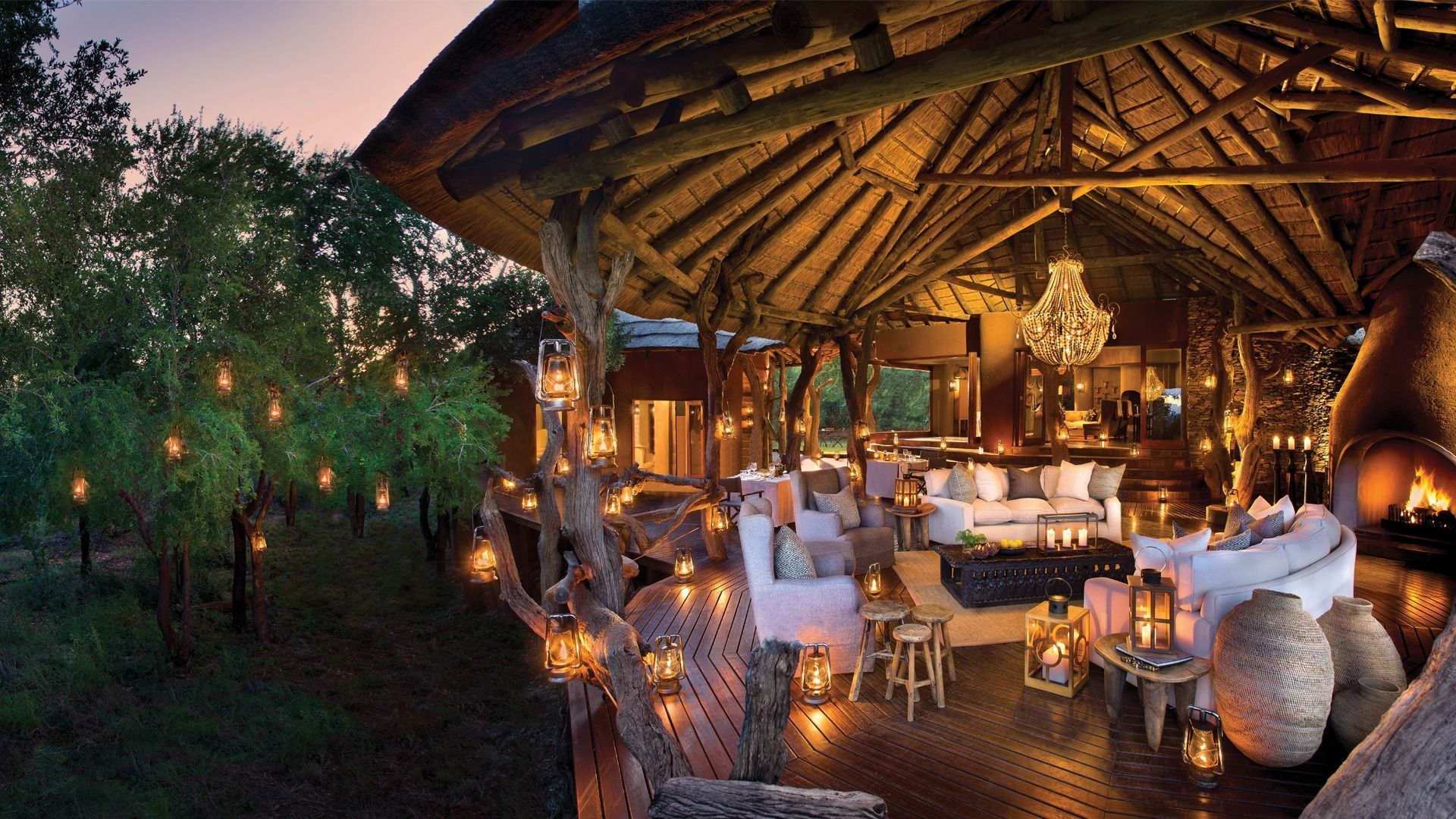 Outdoor lodge with thatched roof, lit by lanterns and strings of lights, with seating and a fireplace.