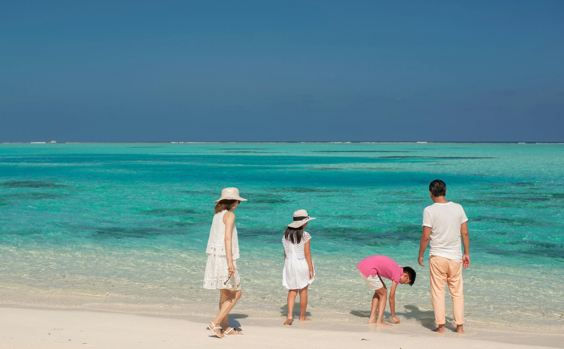 Family exploring a turquoise beach. Person in pink shirt bends down; others walk or stand, looking at the water. Sunny day. Maldives Family Holiday