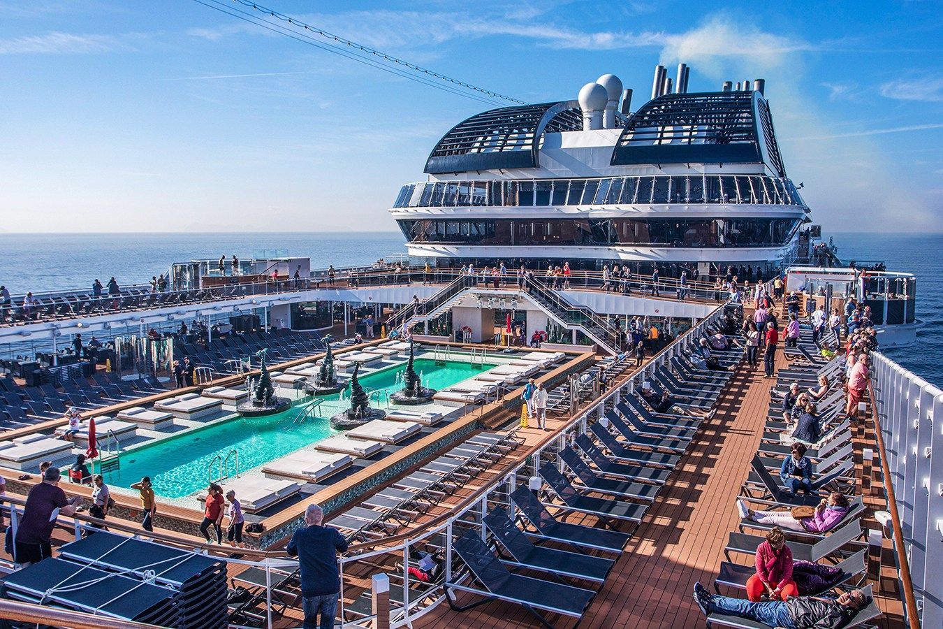 Cruise ship deck with pool, sunbathers, and multiple decks overlooking ocean.