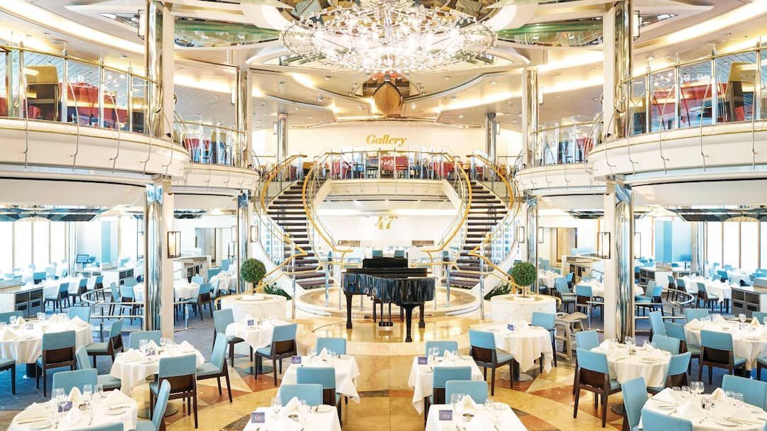 Grand dining room with white tables, blue chairs, a grand piano, and a dual staircase on a cruise ship.