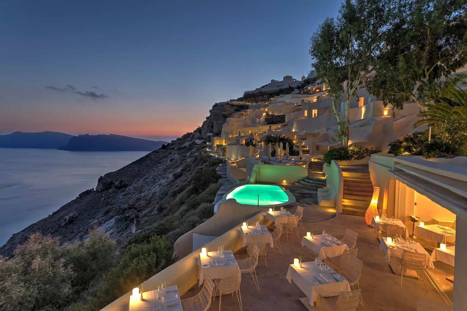 Restaurant on a cliff overlooking the sea at dusk; tables set for dining, lit pool, white buildings. Mystique, a Luxury Collection Hotel