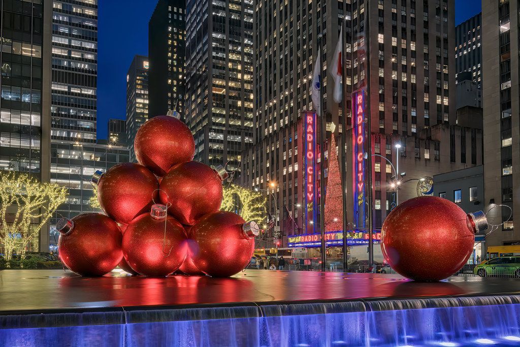 Large red Christmas ornaments on a fountain, Rockefeller Center at night. Tall buildings and Radio City in the background.