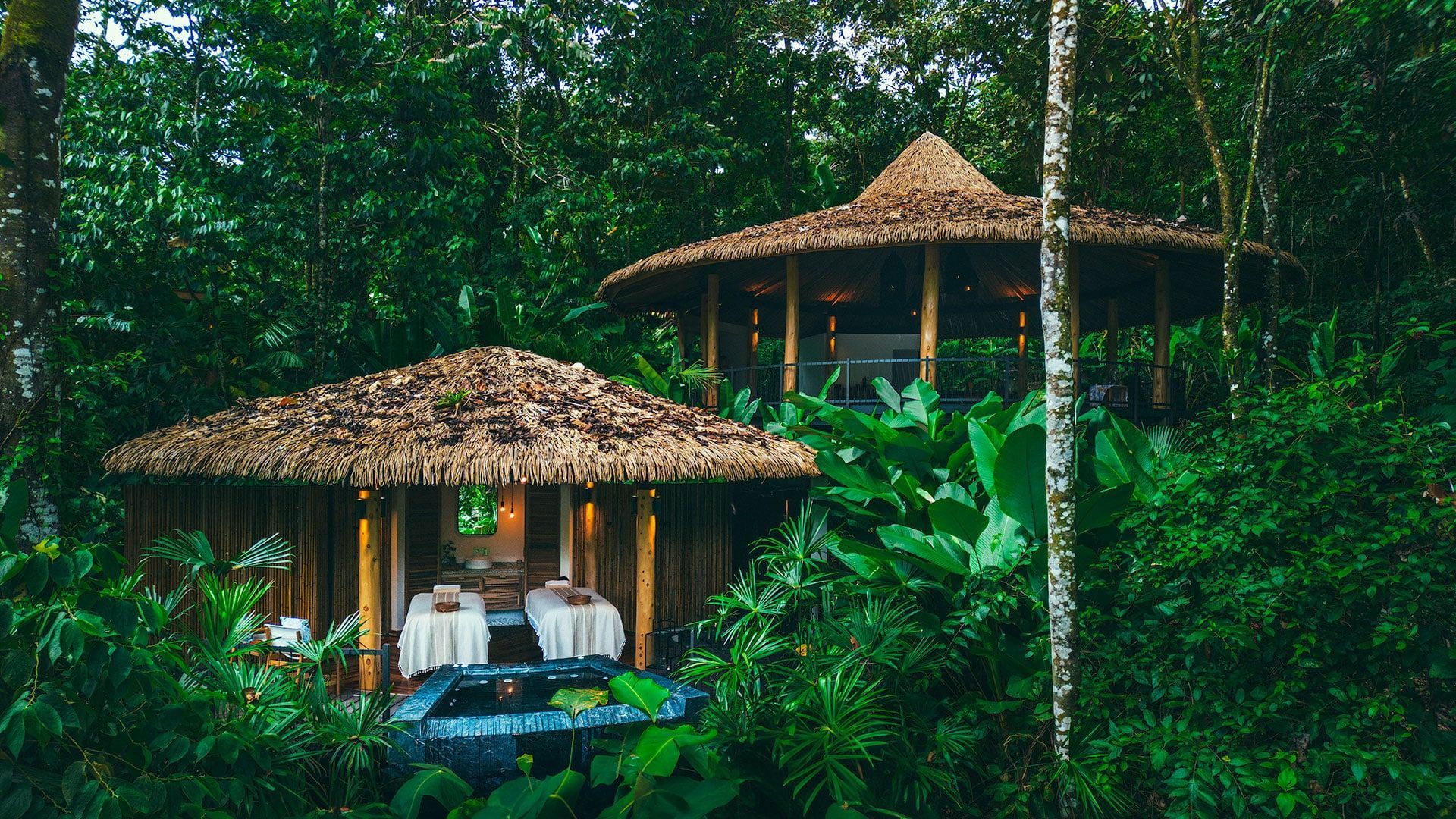 Two thatch-roofed huts in lush green jungle, likely a spa setting.