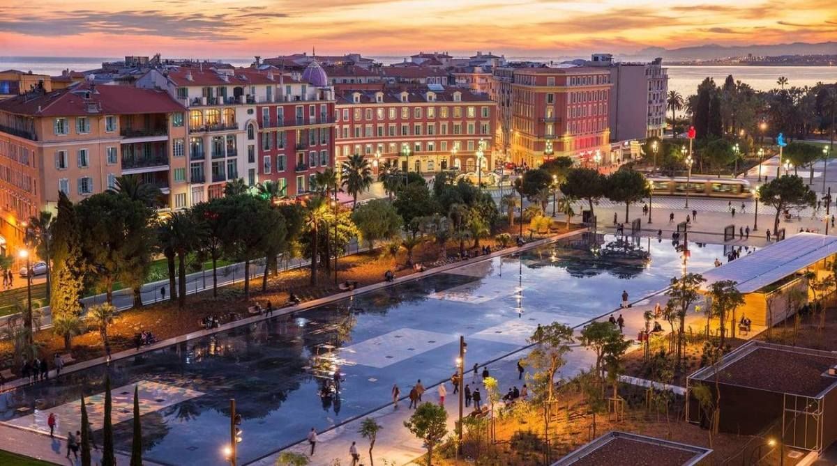 Evening view of a city square with illuminated buildings, reflecting pool, trees, and people; Nice, France.