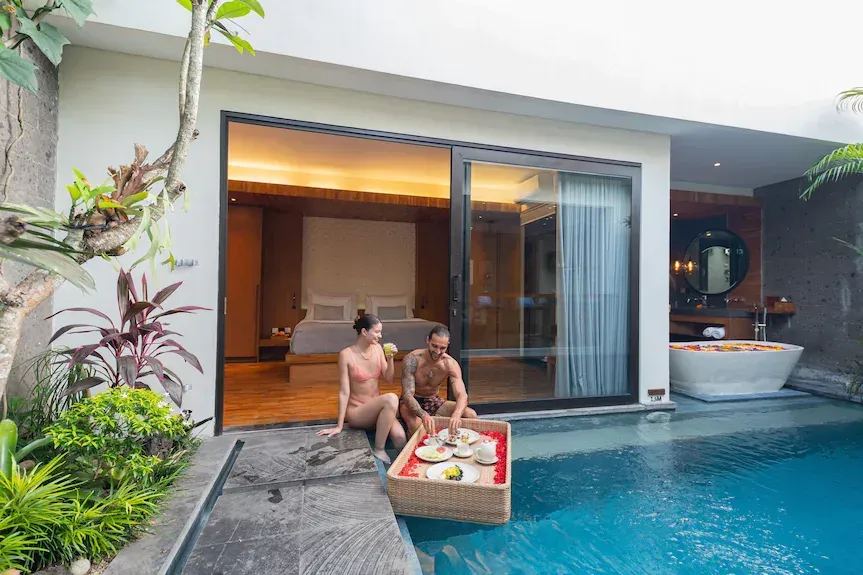 Couple in swimwear enjoying floating breakfast in private pool by a room with open sliding glass doors. Nusa Dua