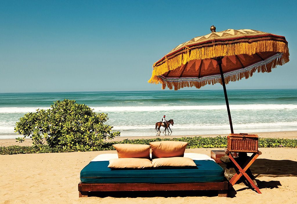 Beach scene with a daybed, ornate umbrella, and ocean view; a person on a horse is in the water. Oberoi, Bali