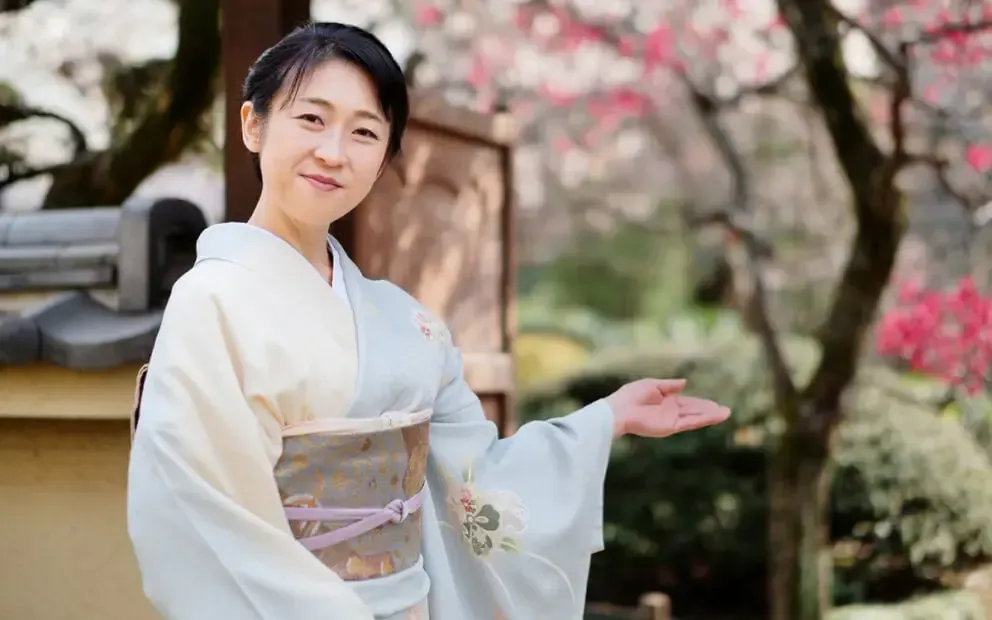 Woman in kimono gesturing toward blooming trees. Omotenashi