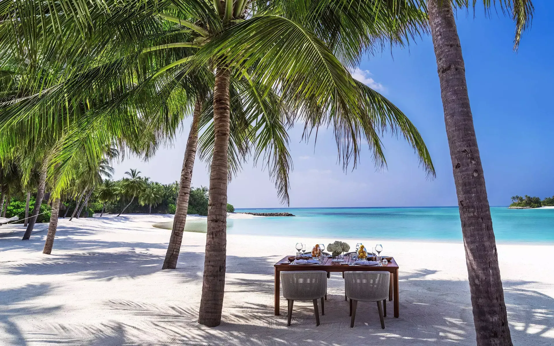 Beach table set for two under palm trees, overlooking turquoise ocean. One & Only Reethi Rah