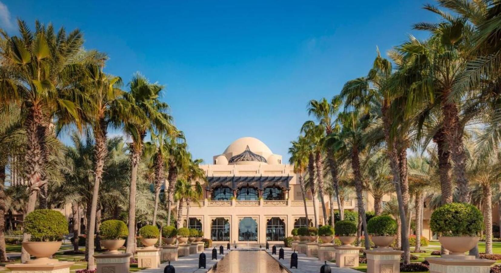 Luxurious beige building with a dome, palm trees, and reflecting pool under a blue sky.