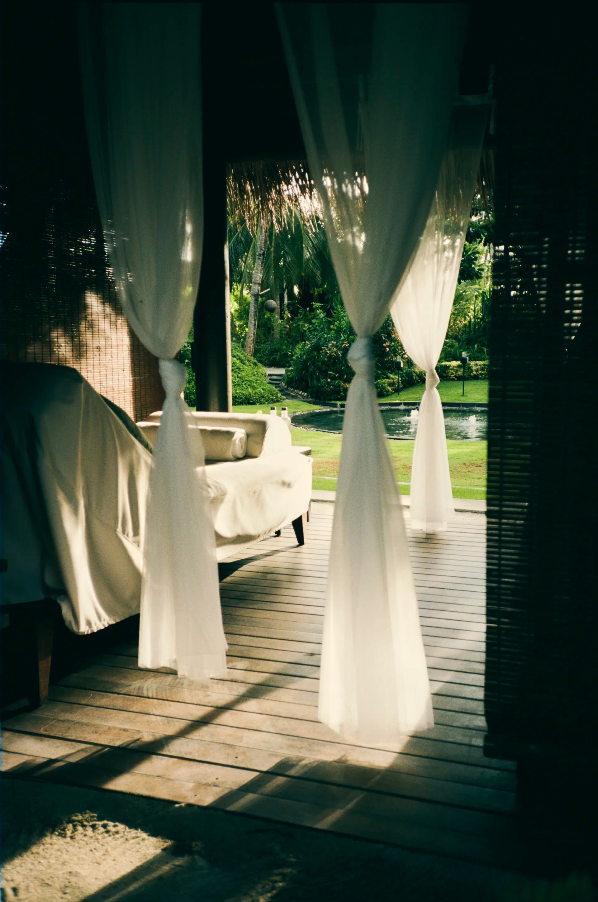 Sheer white curtains tied back, framing a view of a covered car, lawn, and trees. Sunlight streams through. One&Only Reethi Rah Maldives Spa