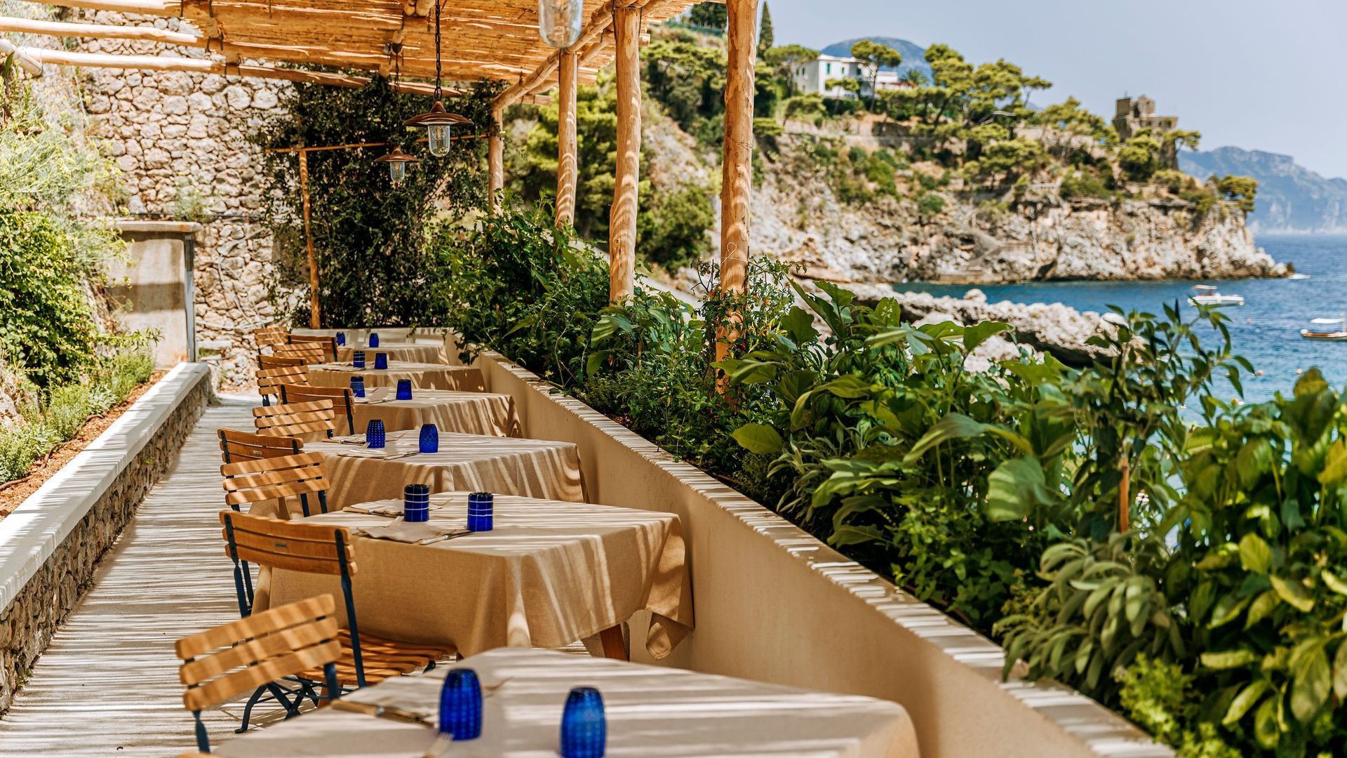 Tables set for outdoor dining on a terrace overlooking the sea, shaded by a wooden pergola and lush green plants. Borgo Santandrea