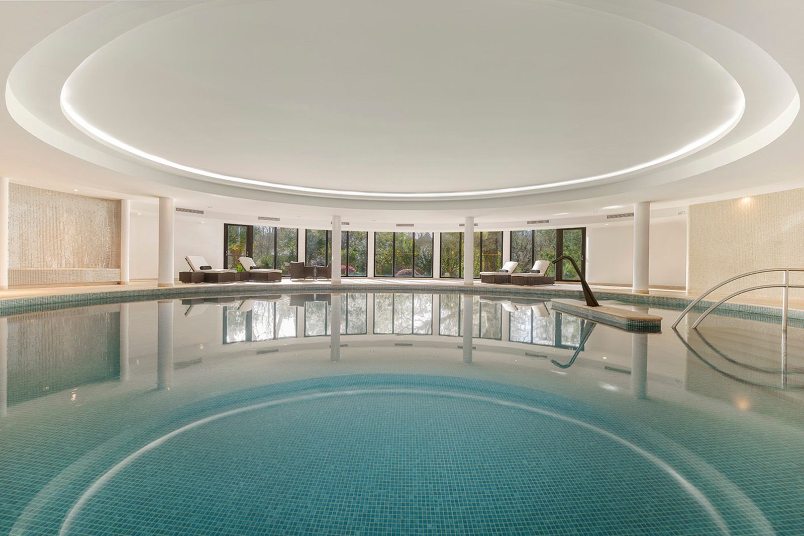 An indoor circular swimming pool under a recessed, lit ceiling, featuring tile details and lounge chairs in the distance.