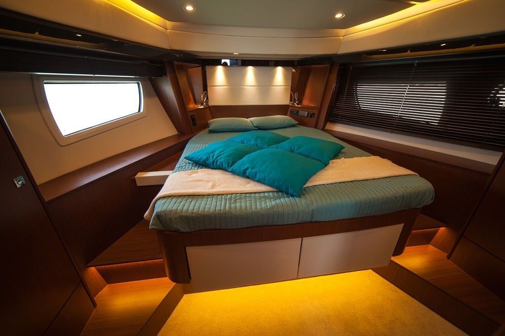 Bedroom interior of a yacht with a bed, turquoise pillows, wooden panels, and porthole window.