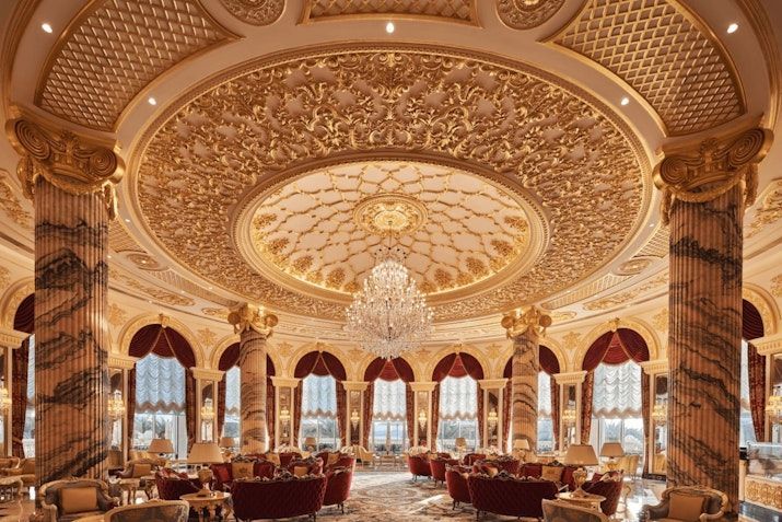 Ornate gold-decorated interior with a large chandelier, domed ceiling, and tall columns. Luxurious seating near windows. Raffles The Palm Dubai
