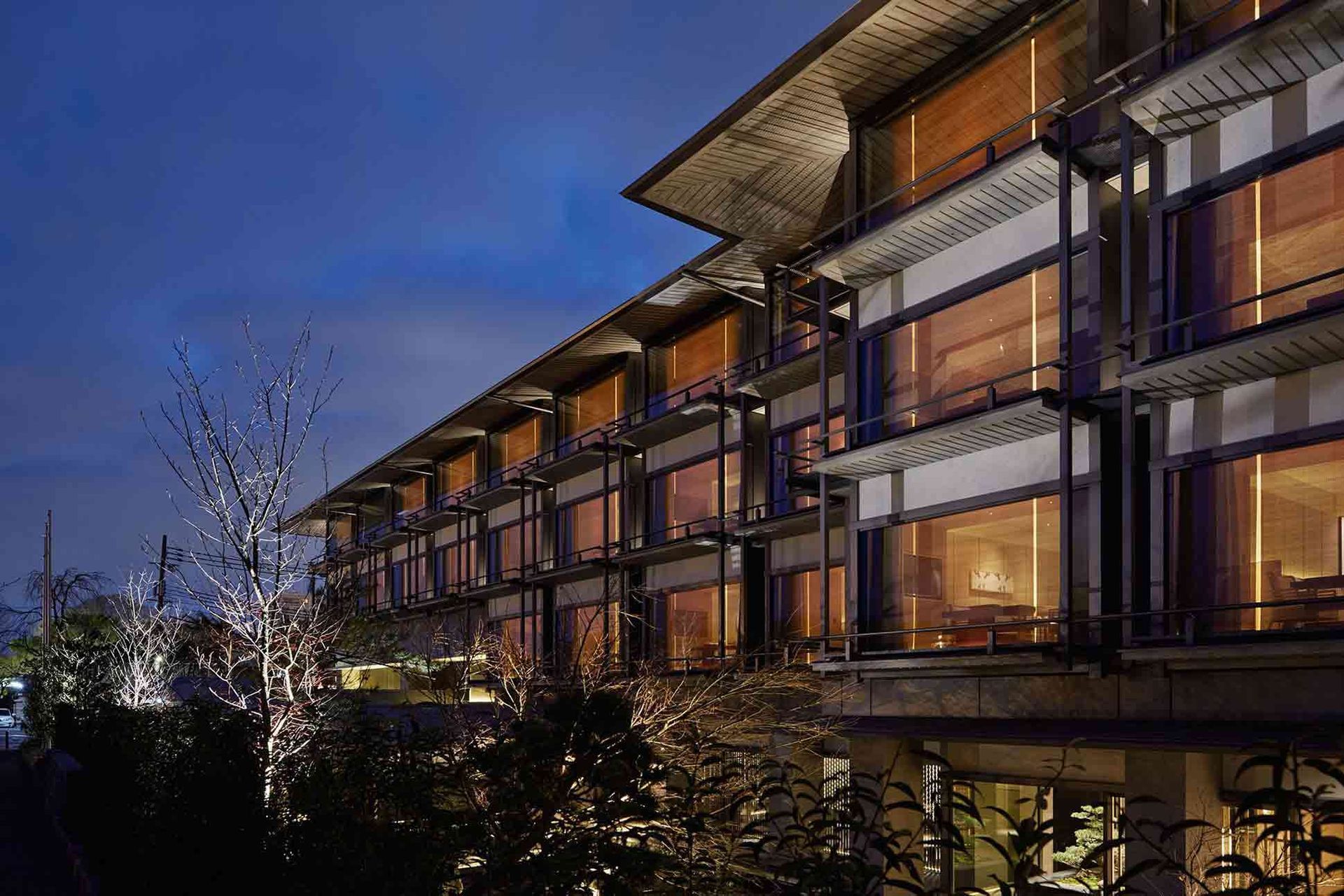 Hotel exterior at dusk, with illuminated rectangular windows and a dark blue sky. Some bare trees are in the foreground. The Ritz-Carlton, Kyoto